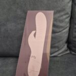 Zafoliowane pudełko Bibi z grafiką wibratora, oparte o szarą kanapę